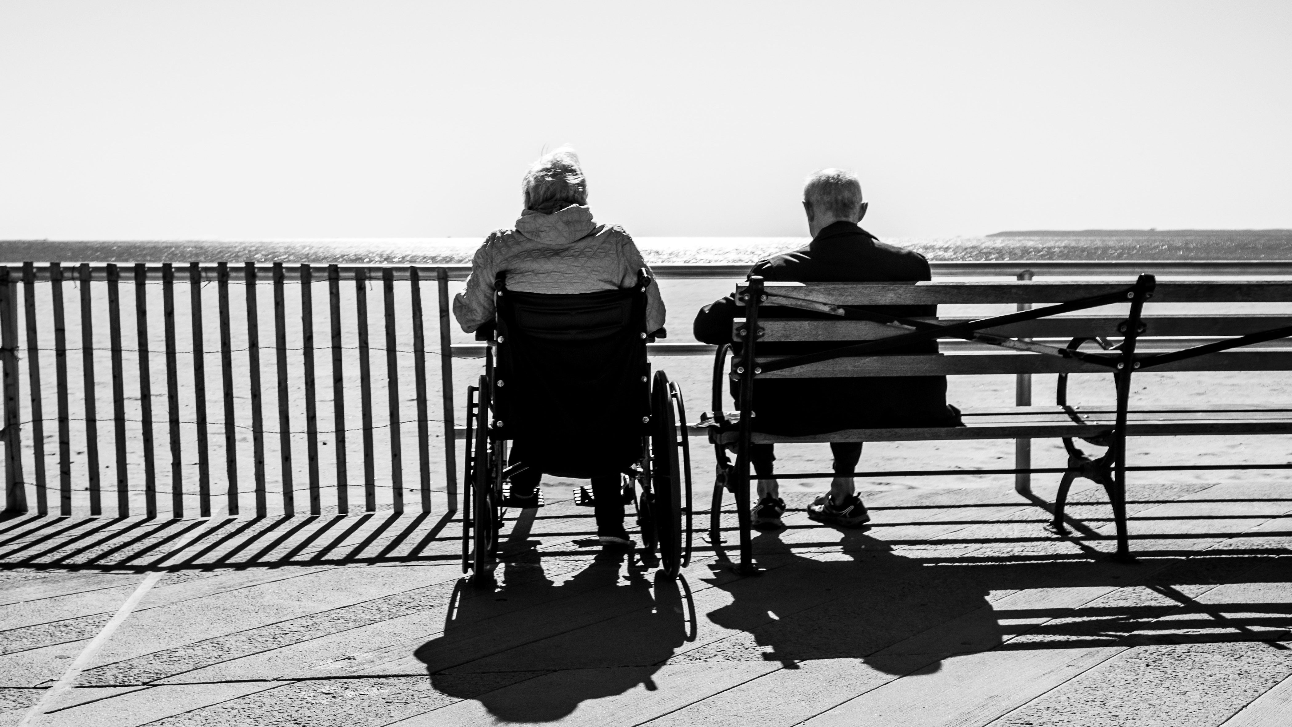 Couple sat together on a promenade by the sea, one in a wheel chair.